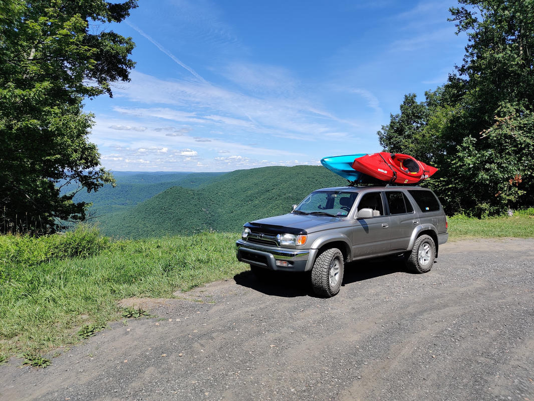 Gray 2001 Toyota 4runner with two kayaks on the roof, parked on a gravel road with a mountainous view